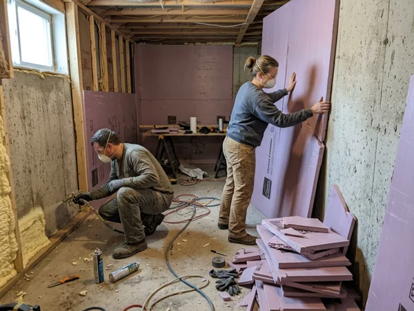 Two tradespeople fitting closed-cell spray foam at the bottom plate and rigid insulation on the foundation wall
